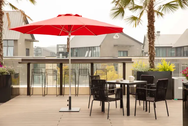 A large red Bossa Nova center-pole market umbrella providing shade for a high-top table and a dining set on a modern wooden patio with palm trees.