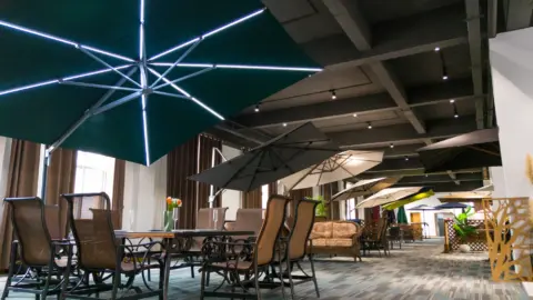 A spacious and elegant showroom displaying a wide range of cantilever umbrellas, including a green model with integrated LED lighting, arranged over stylish outdoor furniture sets.