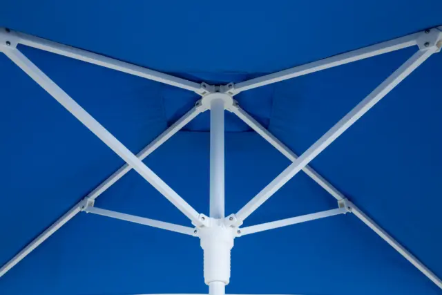 An upward view of the open Frappe center-pole umbrella, highlighting the robust white central pole and support rib structure underneath the vibrant blue canopy.