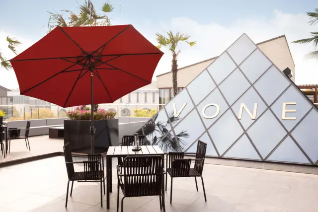 A large, open, red Frappe center-pole umbrella providing shade for a table and chairs on a modern rooftop patio with a glass pyramid structure in the background.