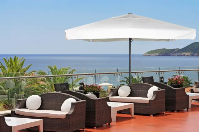 A large white Luciano patio umbrella providing elegant shade for wicker lounge furniture on a beautiful hotel terrace overlooking the ocean.