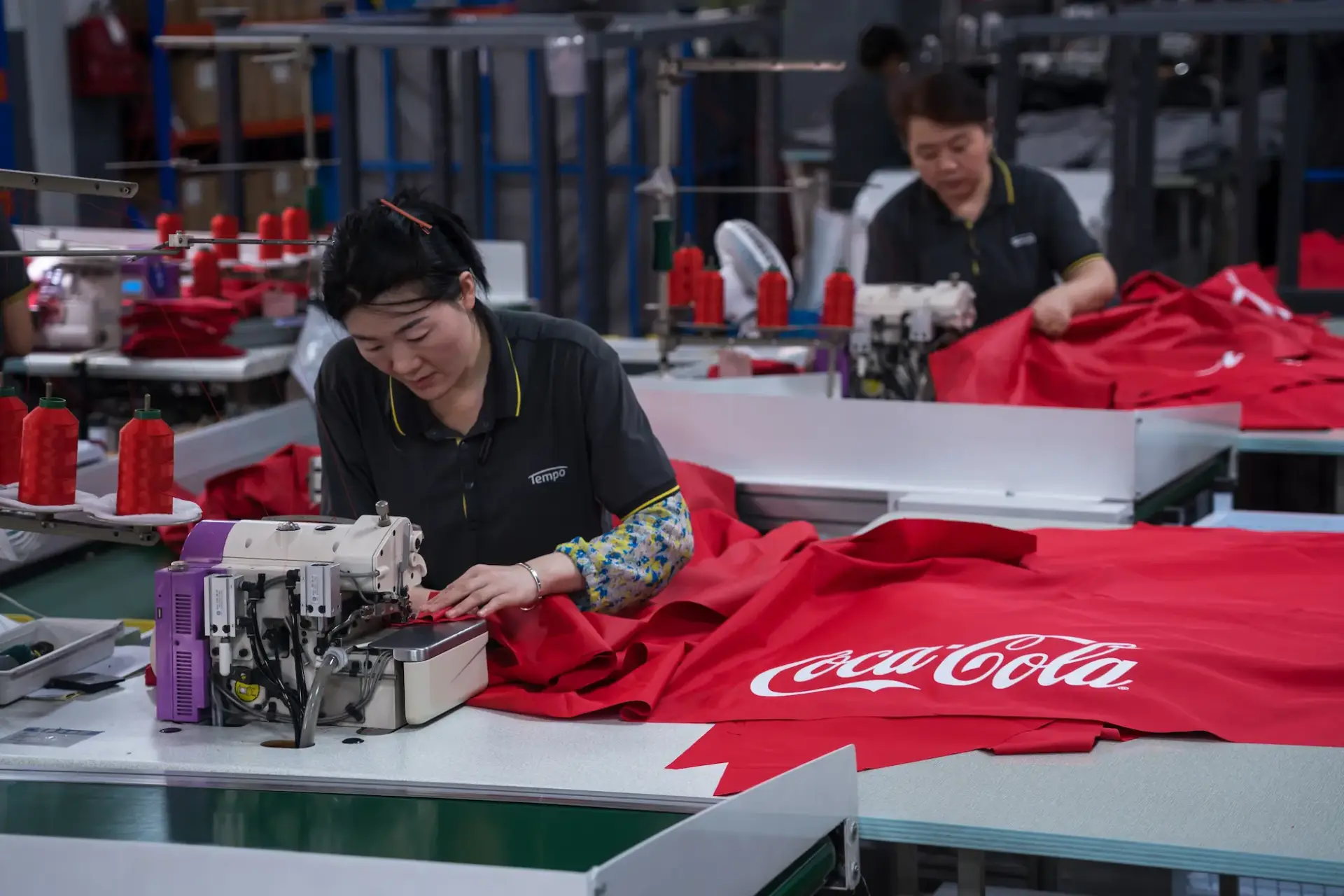 A skilled worker at Tempo Manufacturing operating an industrial sewing machine to construct a vibrant red umbrella canopy with a custom-printed Coca-Cola logo.